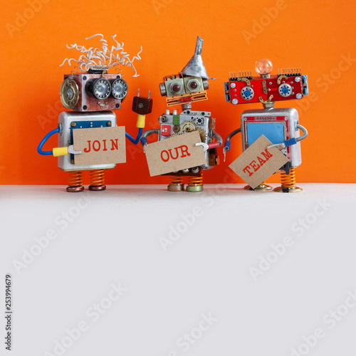 Robotic hiring recruitment concept. Three funny robots looking for a new assistant in company. Hr cyborgs holds a cardboard posters handwritten text Join Our Team. Orange gray background, copy space.