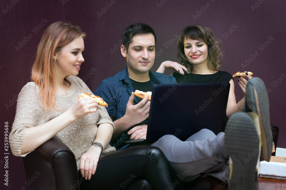 Young people eating pizza at the laptop. A man and two women are watching a movie on a computer. Young people eating pizza at the laptop. A man and two women are watching a movie on a computer.