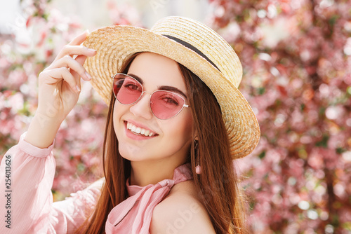  Outdoor close up portrait of young beautiful happy smiling lady wearing stylish pink cat eye sunglasses, straw hat, earrings, blouse, posing in street of city. Copy, empty space for text