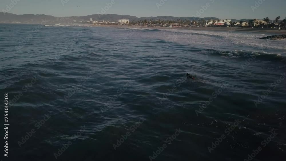 Aerial view behind surfer moving around on ocean waves off the coast of Venice beach. Coastal property to right along skyline. Aerial view behind surfer moving around on ocean waves off the coast of Venice beach. Coastal property to right along skyline.