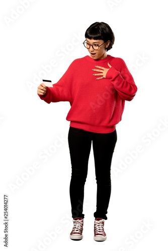 A full-length shot of a Short hair girl with red sweater holding a credit card and surprised over isolated white background