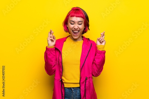 Young woman with pink hair over yellow wall with fingers crossing