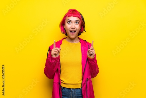 Young woman with pink hair over yellow wall surprised and pointing up