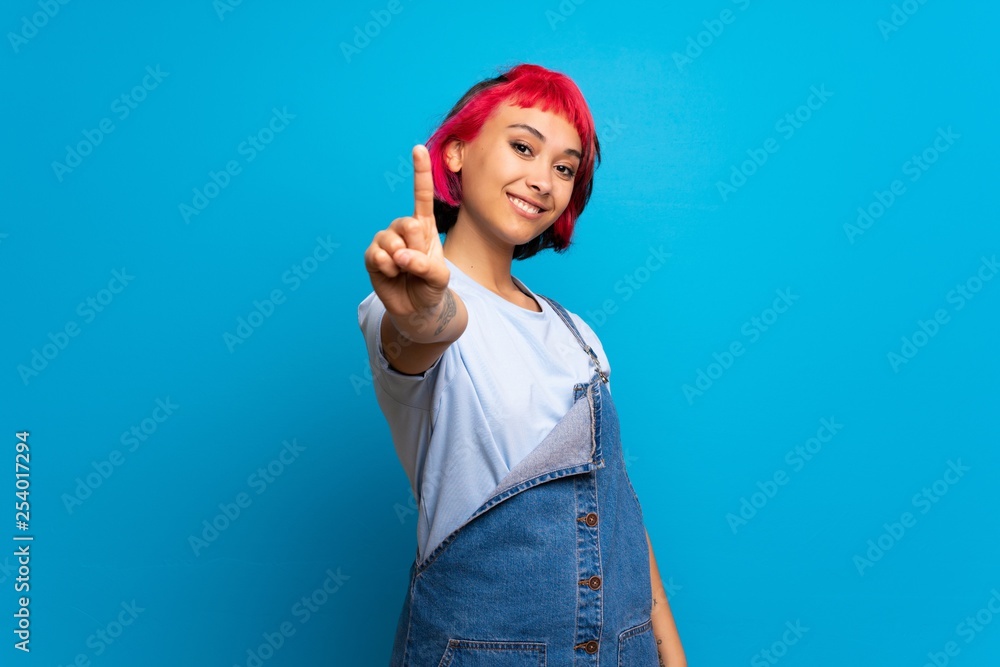 Young woman with pink hair over blue wall showing and lifting a finger Young woman with pink hair over blue wall showing and lifting a finger
