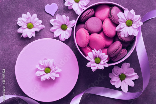 Purple and pink macaroons in a gift box on a beautiful purple background decorated with flowers. Top view