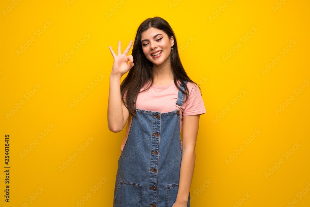 Teenager girl over yellow wall showing ok sign with fingers Teenager girl over yellow wall showing ok sign with fingers