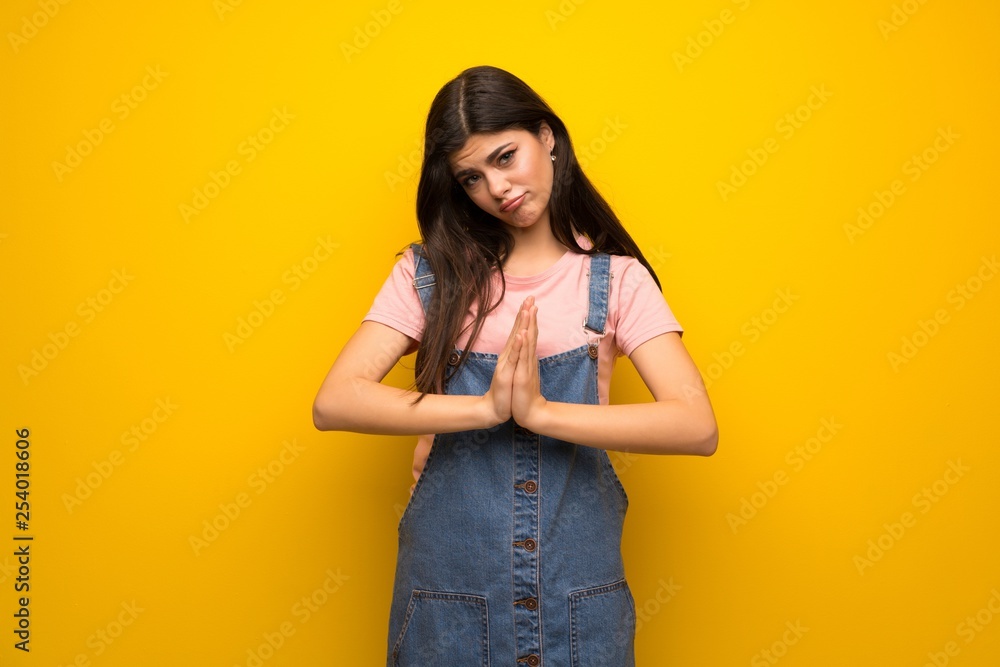 Teenager girl over yellow wall keeps palm together. Person asks for something Teenager girl over yellow wall keeps palm together. Person asks for something