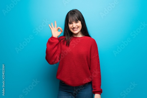 Woman with red sweater over blue wall showing ok sign with fingers