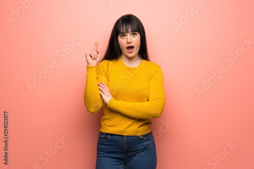 Woman with yellow sweater over pink wall thinking an idea pointing the finger up
