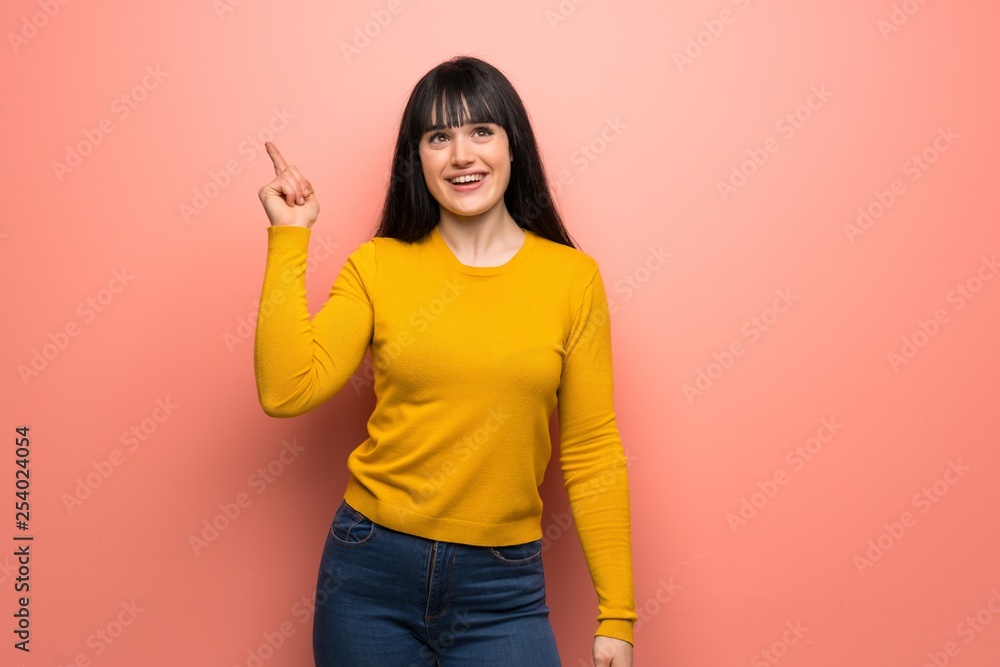 Woman with yellow sweater over pink wall intending to realizes the solution while lifting a finger up Woman with yellow sweater over pink wall intending to realizes the solution while lifting a finger up