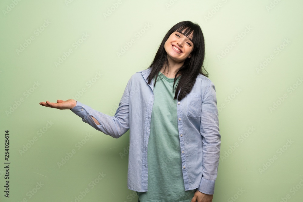 Young woman over green wall presenting an idea while looking smiling towards Young woman over green wall presenting an idea while looking smiling towards