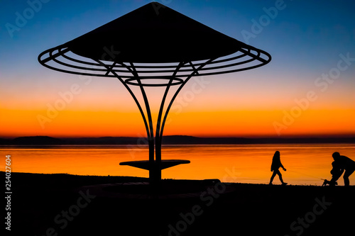 Incredible, bright sunset over the water against the background of which the silhouette of a beach umbrella with a bench and a girl playing with a child and a dog. Minsk Sea, Belarus © Natallia