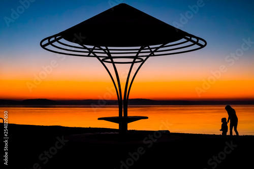 Incredible, bright sunset over the water against the background of which the silhouette of a beach umbrella with a bench, a gazebo. Minsk Sea, Belarus © Natallia