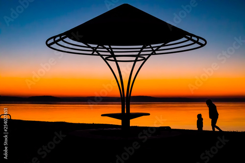 Incredible, bright sunset over the water against the background of which the silhouette of a beach umbrella with a bench and a girl playing with a child and a dog. Minsk Sea, Belarus © Natallia