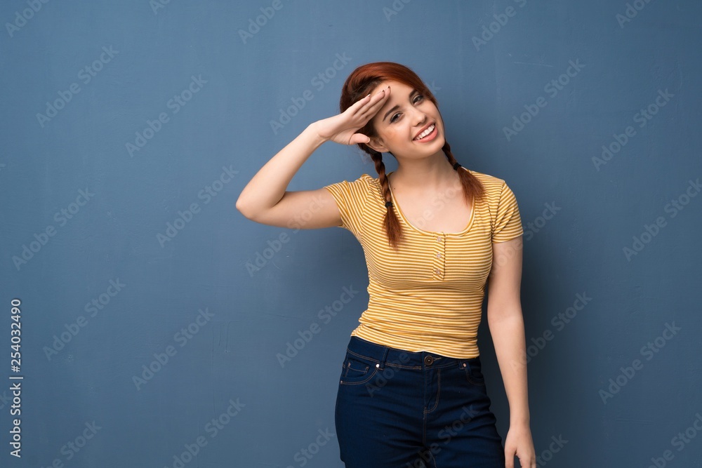 Young redhead woman over blue background saluting with hand Young redhead woman over blue background saluting with hand