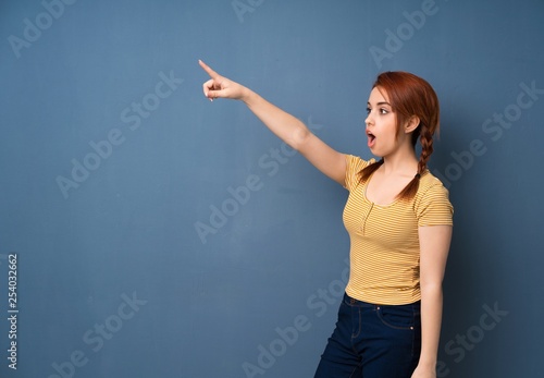 Young redhead woman over blue background pointing away