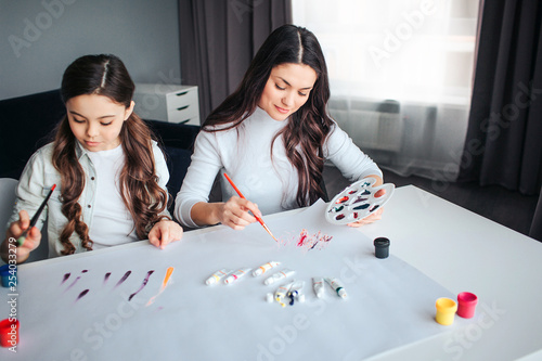Beautiful brunette caucasian mother and daughter paint together in room. They sit at table and draw. Calm cocentrated workers. photo