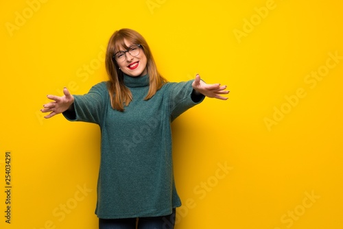 Woman with glasses over yellow wall presenting and inviting to come with hand