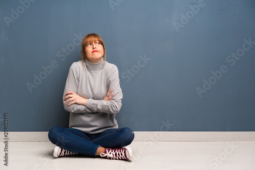 Redhead woman siting on the floor making doubts gesture while lifting the shoulders