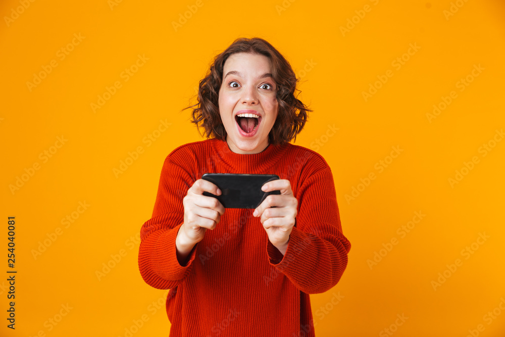 Emotional young pretty woman posing isolated over yellow wall background play games by mobile phone. Emotional young pretty woman posing isolated over yellow wall background play games by mobile phone.