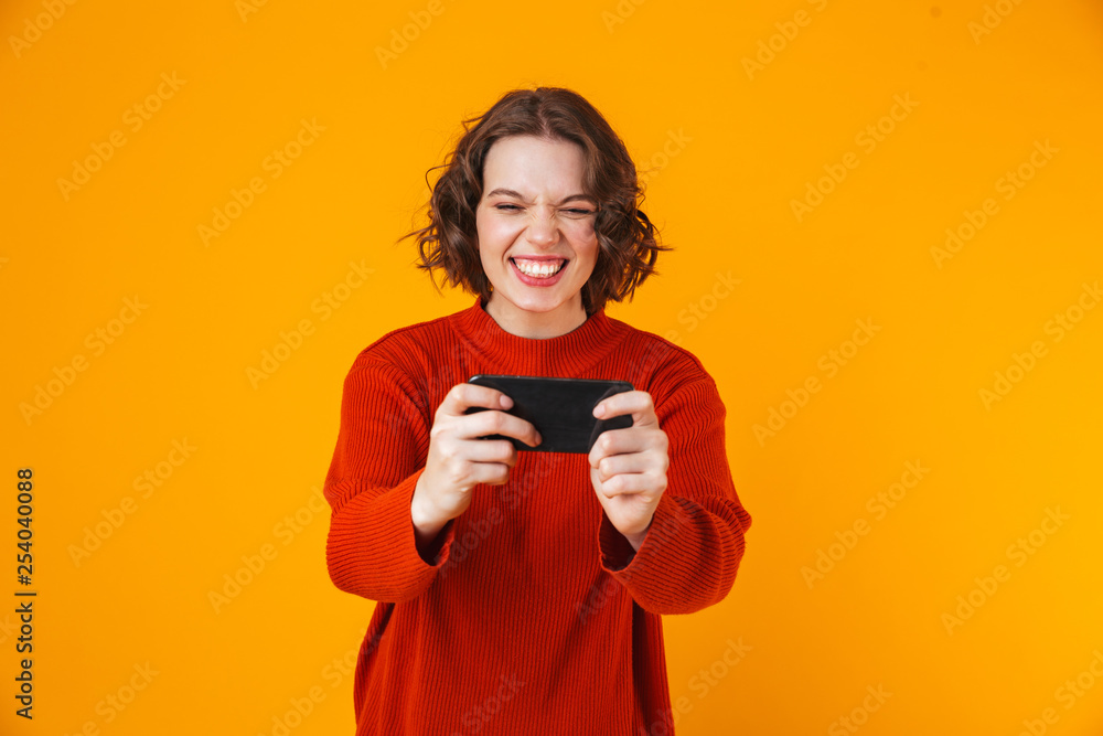 Emotional young pretty woman posing isolated over yellow wall background play games by mobile phone. Emotional young pretty woman posing isolated over yellow wall background play games by mobile phone.
