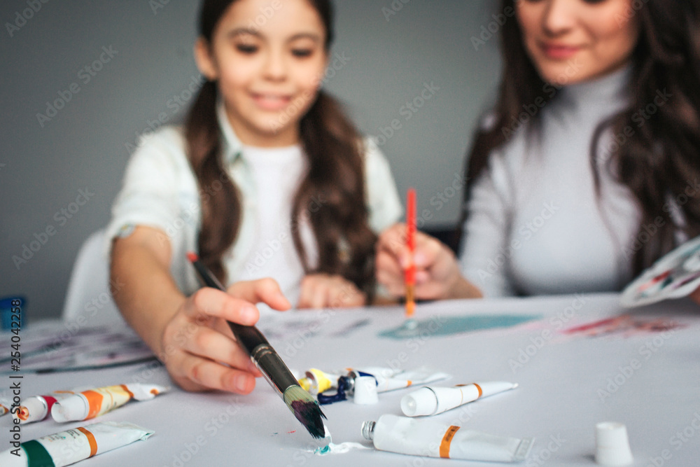 Fototapeta premium Beautiful brunette caucasian mother and daughter paint together in room. Cut view and blurred picture. Happy girl put brush into color. Mother sit besides. Tubes with paint.