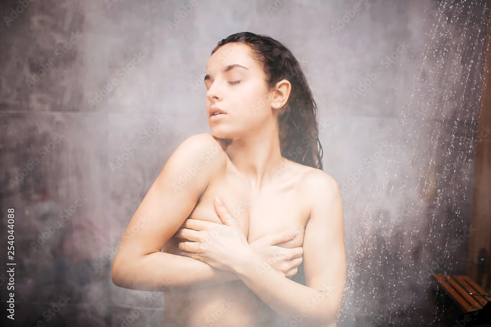 Young attractive sexy woman in shower. Enjoyment during washing herself.  Covering breast with hands. Eyes closed. Water vapor on glass wall. Semi  blurred picture. Photos