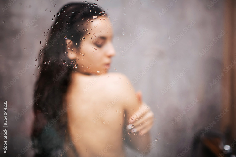 Young attractive sexy woman in shower. Standing back to camera. Embrace herself with closed eyes. Blurred photo. Water vapor on glass wall.