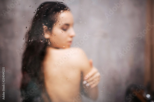 Young attractive sexy woman in shower. Standing back to camera. Embrace herself with closed eyes. Blurred photo. Water vapor on glass wall.