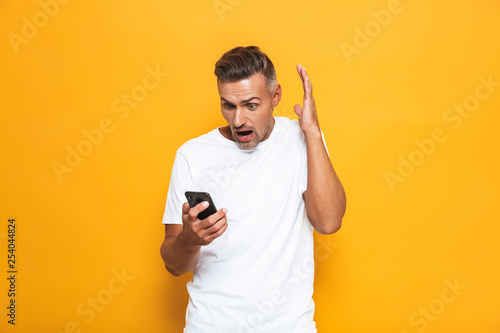 Image of annoyed man 30s in white t-shirt screaming and holding smartphone while standing isolated