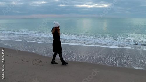 An adult woman walks along a sandy beach at the edge of the water against the backdrop of calm sea. Autumn mood.