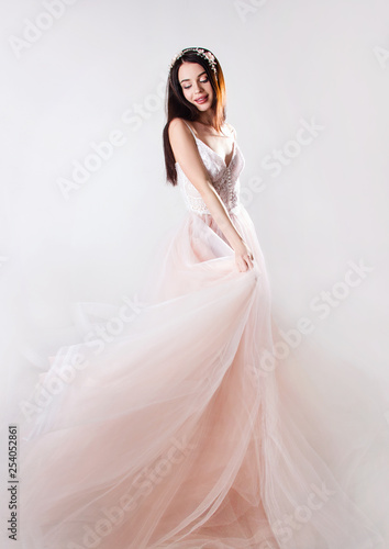 young brunette sexy girl in a white wedding boudoir dress and jewellery in hairstyle is standing and smiling with dress in her hand on a white wall background , spring, bride concept