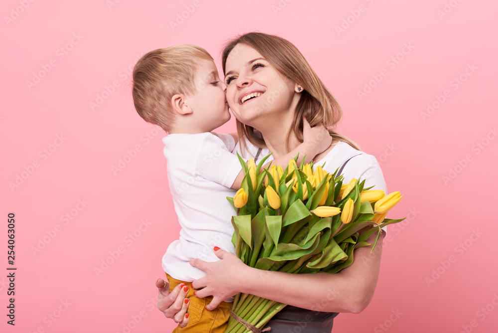 Young mother hugs his little son sitting on the floor against a pink background. Mom holds a bouquet of spring yellow flowers. Care and relationships and family concept. Mothers day