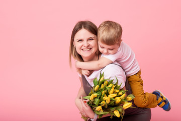 Young mother hugs his little son sitting on the floor against a pink background. Mom holds a bouquet of spring yellow flowers. Care and relationships and family concept. Mothers day
