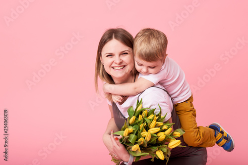 Obraz na płótnie Young mother hugs his little son sitting on the floor against a pink background. Mom holds a bouquet of spring yellow flowers. Care and relationships and family concept. Mothers day w salonie