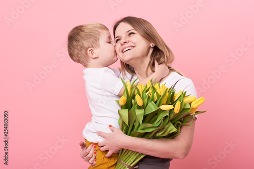 Obraz na płótnie Young mother hugs his little son sitting on the floor against a pink background. Mom holds a bouquet of spring yellow flowers. Care and relationships and family concept. Mothers day w salonie