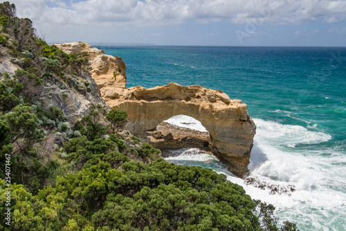 A sea arch on the Great Ocean Road in Victoria, Australia. There are plants in the foreground, a blue/green ocean, and blue sky with clouds.
