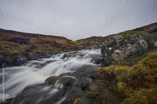 Wild stream or creek in moorland or heath in the highland of Faroe island Vagar with waterfall cascades and stones with moss and lichen on them. Picture taken in cloudy, misty, foggy and rainy day.