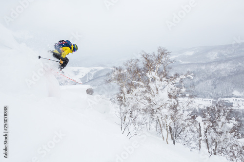 Man in yellow jumps off a cornice near the summit of a small peak in the backcountry of Hokkaido, Japan.