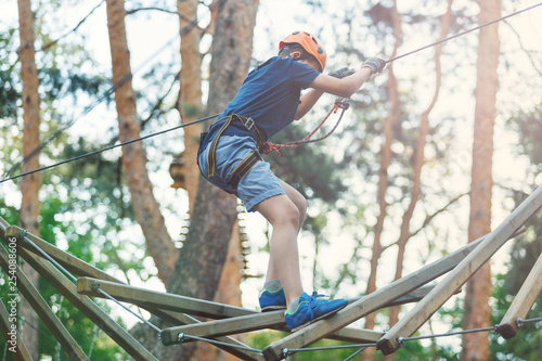 Sporty, young, cute boy in white t shirt spends his time in adventure rope park in helmet and safe equipment in the park in the summer. Active lifestyle concept