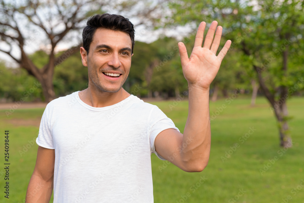 Happy young handsome Hispanic man waving hand at the park Happy young handsome Hispanic man waving hand at the park