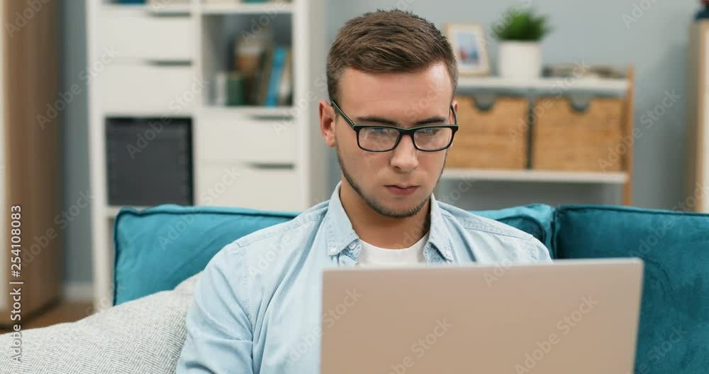 Close up of the young handsome Caucasian guy in glasses doing something on the laptop computer and then smiling to the camera while sitting at home on the couch. Portrait. Close up of the young handsome Caucasian guy in glasses doing something on the laptop computer and then smiling to the camera while sitting at home on the couch. Portrait.