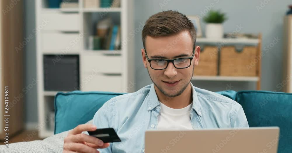 Portrait of the young Caucasian man in glasses sitting on the couch in the living room and being very cheerful because buying online with a credit card on the laptop computer. Close up. Portrait of the young Caucasian man in glasses sitting on the couch in the living room and being very cheerful because buying online with a credit card on the laptop computer. Close up.