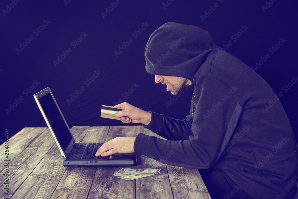 hacker in black hoody with laptop, bank card and dollar notes hacker in black hoody with laptop, bank card and dollar notes