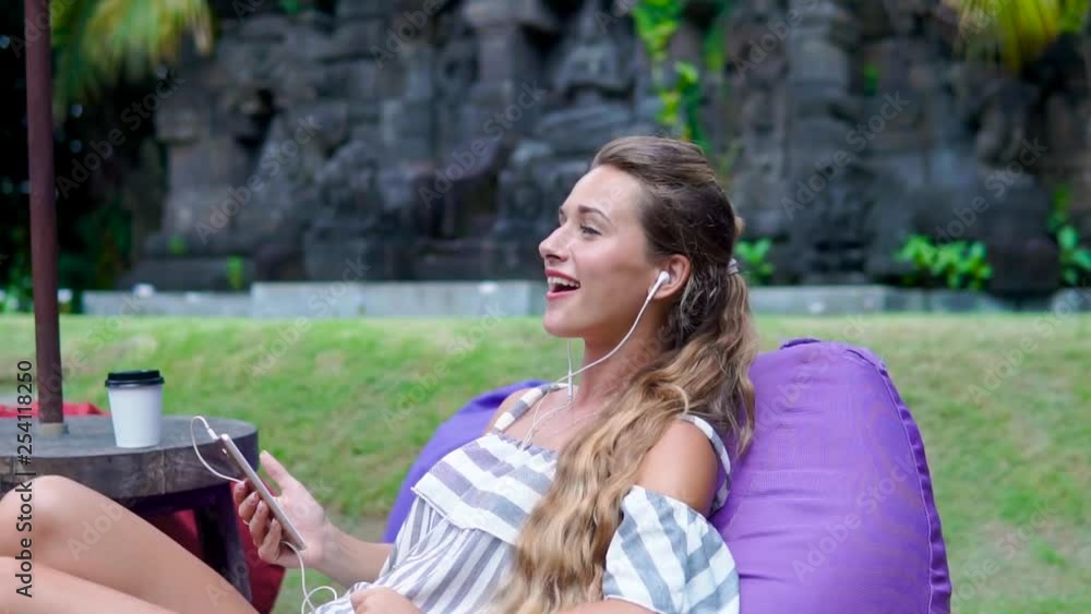 Fashion girl drinks coffe while she is writing a message while sitting on couch in green garden on background of sculpture in Balinese style Fashion girl drinks coffe while she is writing a message while sitting on couch in green garden on background of sculpture in Balinese style