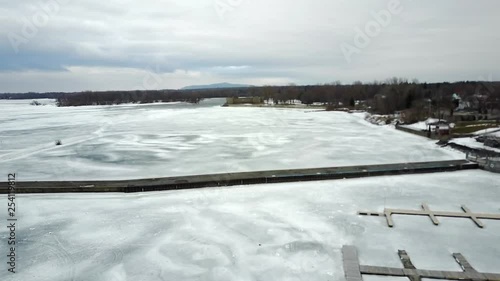Cinematic aerial / drone footage moving forwards over the lake with dry trees, snow, houses, a marina, mountains and a fort in the background in Chambly, Quebec, Canada during winter season. photo