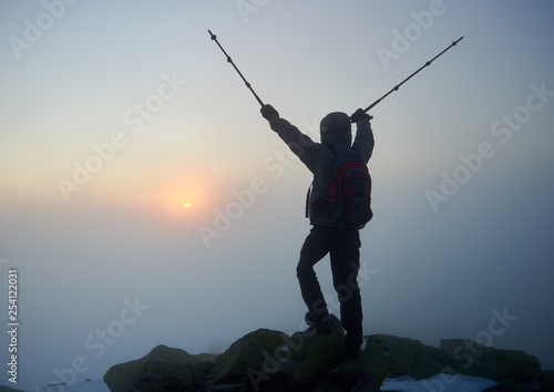 Back view of tourist hiker in warm jacket with backpack on rocky mountain peak with hiking stick in raised arms on copy space background of foggy misty blue sky and raising bright orange sun at dawn.