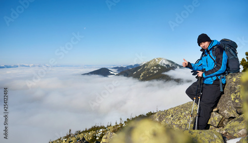 Tourist hiker with backpack and trekking poles smiling in camera on steep rocky mountain slope on background of beautiful blue sky, foggy valley filled with white clouds and snowy mountain tops.