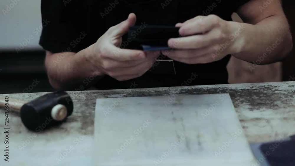 The master of creating leather wallets handmade hammers the product, applies glue with a brush. Close up of a master working with leather textile at his workshop using tools.