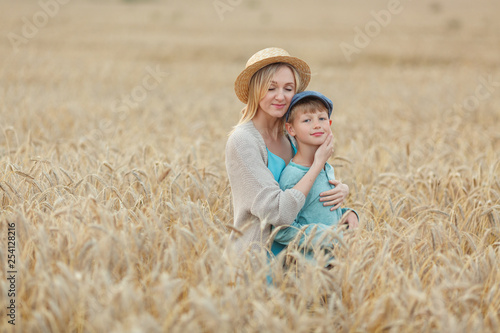 Mom with a straw hat and blue dress hugs her son in a cap standing in a wheat field. Summer holidays in the village © Diana
